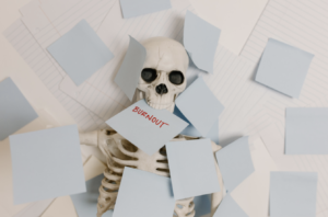 Skeleton buried under sticky notes with the word “burnout,” symbolizing workplace exhaustion and stress.