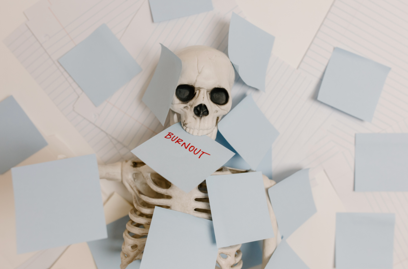 Skeleton buried under sticky notes with the word “burnout,” symbolizing workplace exhaustion and stress.